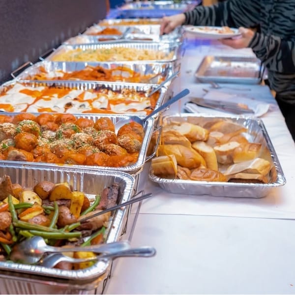 A buffet line with trays of lasagna, meatballs, roasted vegetables, and bread, with a person serving food.