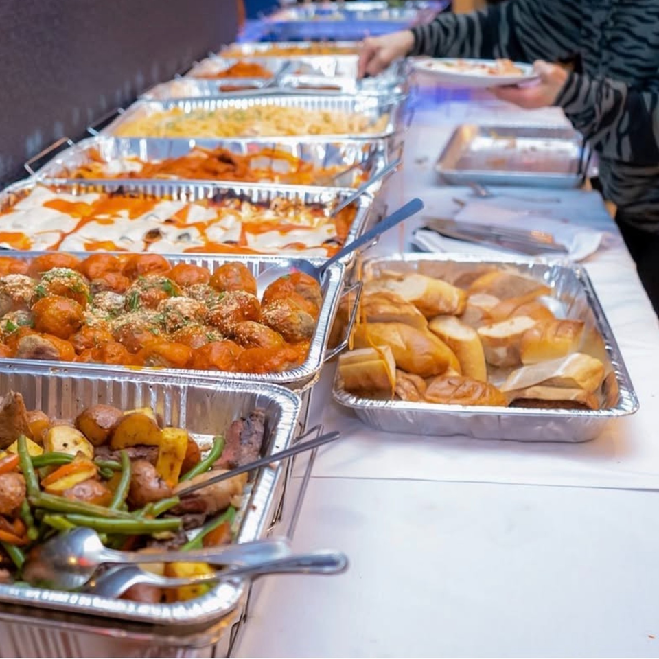 A buffet line with trays of lasagna, meatballs, roasted vegetables, and bread, with a person serving food.