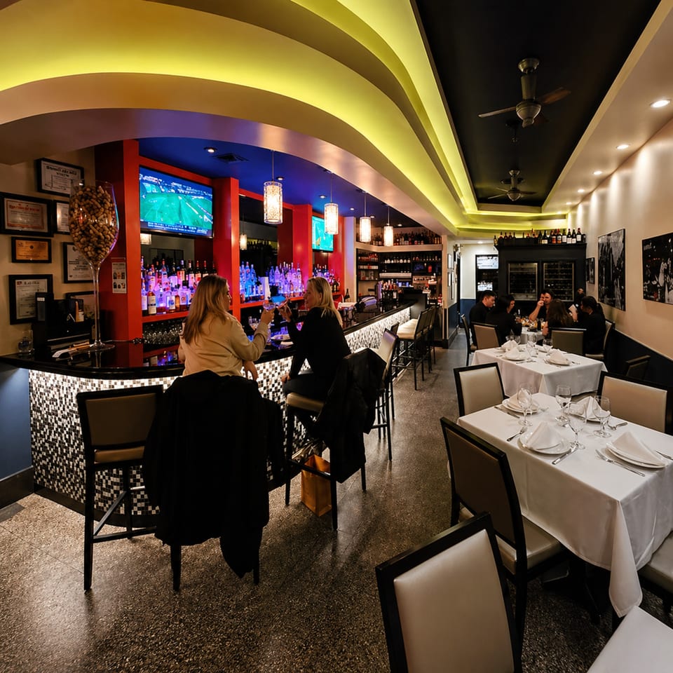 Two women drink at a lively bar with TVs, while other patrons dine at white-clothed tables.
