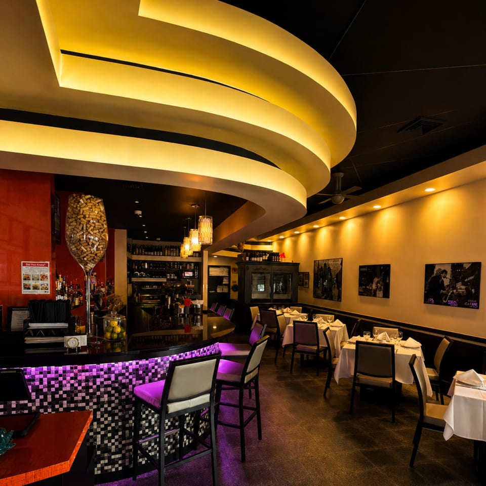 A restaurant interior with a bar featuring purple lighting and dining tables with white tablecloths.