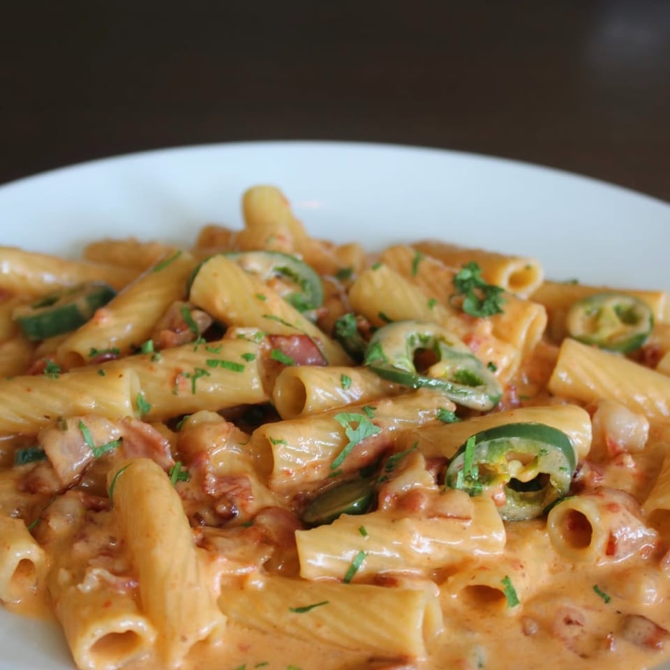 Rigatoni pasta in a creamy orange sauce with bacon bits, jalapeño slices, and parsley on a white plate.