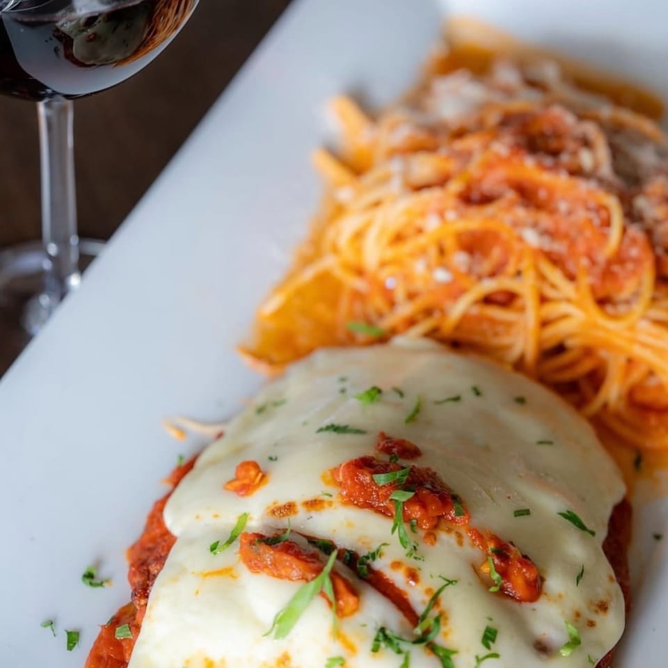 Chicken parmesan with spaghetti and red sauce, topped with cheese and parsley, next to a glass of red wine.
